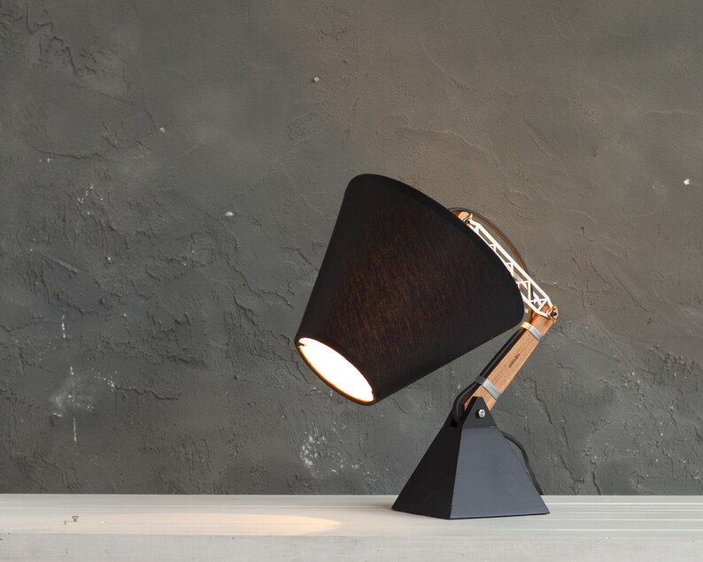 Peut inclure: Une lampe de bureau noire moderne avec un abat-jour conique. La lampe est dot&eacute;e d'un bras en bois et d'une structure m&eacute;tallique g&eacute;om&eacute;trique. La base est de forme pyramidale noire. La lampe est sur une surface blanche contre un mur gris textur&eacute;.