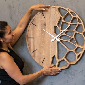 Peut inclure: Une grande horloge murale ronde en bois avec un design unique. L'horloge présente une finition en grain de bois naturel et un design ajouré partiel sur le côté droit. Des lignes noires marquent les heures, et les aiguilles sont blanches.