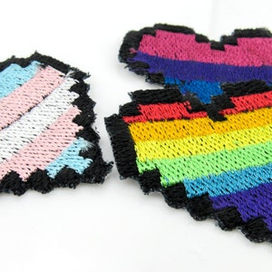 Puede incluir: Parches bordados con forma de corazón en estilo pixelado. Un parche presenta los colores de la bandera transgénero, otro la bandera bisexual y un tercero un diseño de arcoíris. Cada parche tiene un borde negro.