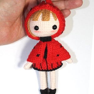 May include: A crocheted doll keychain featuring a character with light skin, brown hair, and black bead eyes. The doll wears a red hooded cape with small black accents, a black collar, a red skirt with black trim, and black boots, attached to a metal ring.