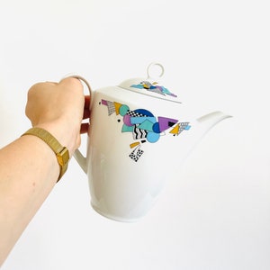 Könnte beinhalten: Eine weiße Teekanne aus Keramik mit einem geometrischen Design in Blautönen, Lila, Gelb und Schwarz. Die Teekanne hat einen Deckel mit einem Ringgriff.