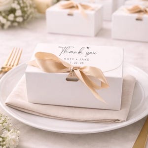 Puede incluir: Caja de regalo blanca con una cinta de raso dorada atada con un lazo. La caja está sobre un plato blanco con una servilleta beige. El texto "Thank you" está impreso, junto con los nombres "KATE + JAKE" y la fecha "7.22.28".