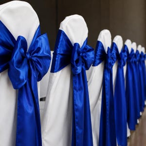 Op de afbeelding: Een rij witte stoelhoezen, elk versierd met een fel koningsblauwe satijnen sjerp die aan de achterkant tot een grote, opvallende strik is geknoopt. De gladde, reflecterende stof van de sjerpen contrasteert met de matte witte hoezen.