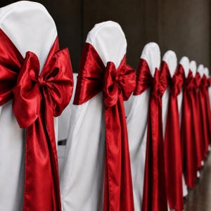 May include: A row of chairs covered in smooth white fabric, each featuring a vibrant red satin sash tied into a large, prominent bow on the backrest. The chairs are arranged in a line, showcasing the consistent white covers and red bows.