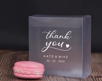 May include: Clear plastic box with a pink macaron inside. The box has a white label with the text "thank you" and a small heart symbol. Below the text is "Kate & Mike" and the date "02.02.2032".