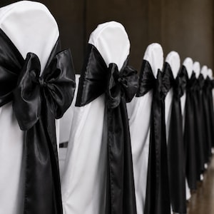 May include: A row of chairs covered in smooth white fabric, each featuring a large, shiny black satin bow tied around the backrest. The fabric covers the entire chair, creating a uniform appearance with the black sashes draped vertically.