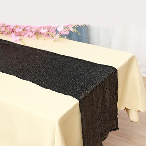Op de afbeelding: Een zwarte pailletten tafelloper ligt op een crèmekleurig tafelkleed. Roze bloemen liggen op de tafel. De loper voegt elegantie toe aan de tafelsetting. De tafel is gedekt voor een speciale gelegenheid. De tafelloper is ongeveer 150 cm lang.