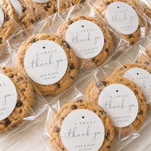 May include: Close-up of several chocolate chip cookies, each individually wrapped in clear cellophane. Each cookie has a white circular sticker with the words "A Sweet Thank You", the names "Jack & Kate" and the date "08.08.2028".