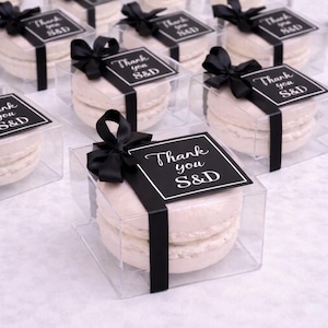 May include: Clear plastic boxes each contain a light-coloured macaron. The boxes are tied with a black ribbon and a black tag that reads "Thank you S&D". The macarons are stacked and appear to be a dessert or treat.