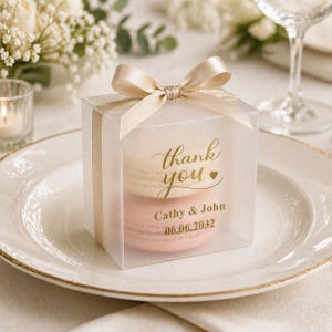 May include: A clear box tied with a gold ribbon, containing two macarons. The box has gold text that reads "thank you" and the names "Cathy & John" with a date. The box sits on a white plate with a gold rim.
