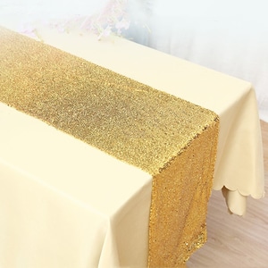 May include: Gold sequin table runner on a white tablecloth. The runner is approximately 14 inches wide and 72 inches long.