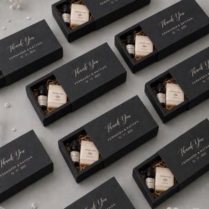 Può includere: Diverse scatole regalo rettangolari nere a scorrimento contenenti una piccola boccetta in vetro ambrato e una saponetta rettangolare naturale adagiata su carta marrone. La fascetta riporta il testo stampato in bianco Thank You, FERNANDA & NATHAN, 12.17.2023.