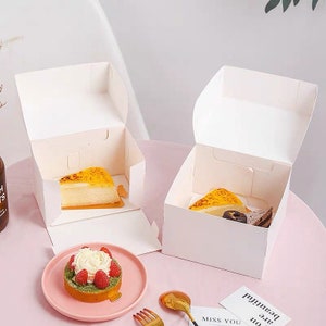 May include: Two open white cardboard bakery boxes each contain a slice of cake. A pink plate holds a small tart topped with cream and raspberries. A gold spoon and fork are next to a card that says "Miss You".