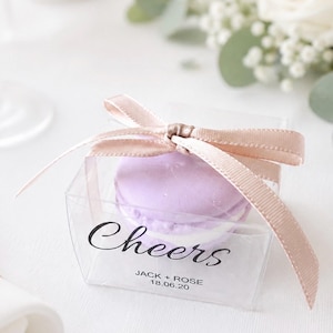 May include: A clear, square box tied with a light pink ribbon, containing a lavender macaron. The box has the word "Cheers" printed on it in black script, along with the names "Jack + Rose" and the date "18.06.20".