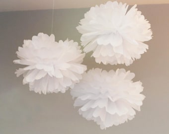 May include: Three large, fluffy white paper pom-poms suspended from the ceiling. Each pom-pom is round with multiple layers of delicate, ruffled paper, creating a soft, cloud-like appearance. Ideal for party decorations.