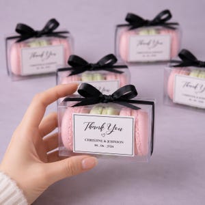May include: Clear plastic favour boxes containing pink and light green macarons, each tied with a black satin ribbon. A white rectangular label on the front features the text Thank You, the names Christine & Johnson, and the date 06.06.2026.