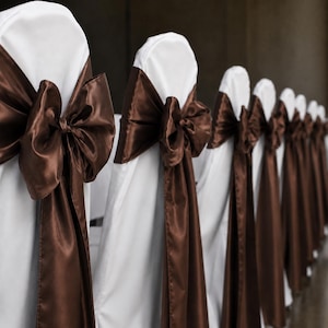 Puede incluir: Una fila de fundas blancas para sillas, cada una con una banda de satén marrón oscuro lisa atada en un lazo grande y prominente en el respaldo. La tela cubre la longitud de las sillas, creando una apariencia uniforme y decorativa en línea.