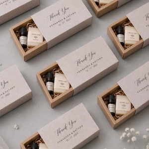 May include: Multiple rectangular cardboard gift boxes containing a small amber glass essential oil bottle and a rectangular bar of natural soap nestled in brown paper filler. The sliding sleeve is printed with Thank You, Fernanda & Nathan, 12.17.2023.