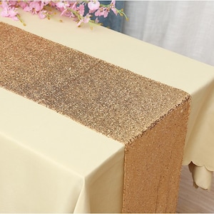 Könnte beinhalten: Ein goldfarbener, mit Pailletten besetzter Tischläufer auf einer cremefarbenen Tischdecke. Der Läufer ist etwa 35 cm breit und 183 cm lang.