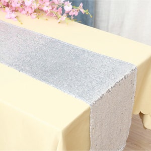 May include: A silver sequin table runner is centered on a pale yellow tablecloth. Pink flowers are in the upper left corner. The runner adds a touch of sparkle to the table setting. The runner is approximately 12 inches wide.