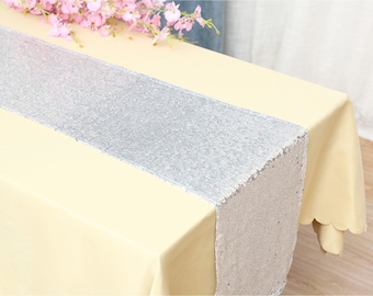 Puede incluir: Un camino de mesa plateado con lentejuelas está centrado en un mantel amarillo pálido. Hay flores rosas en la esquina superior izquierda. El camino de mesa añade un toque de brillo. El camino de mesa mide unos 30 cm de ancho.