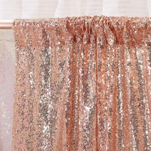 May include: A rose gold sequin fabric backdrop featuring a dense arrangement of small, reflective, circular sequins covering the entire surface. The material is gathered at the top, creating vertical folds and a shimmering, textured appearance.