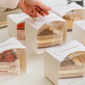 Könnte beinhalten: Weiße Pappkartons mit durchsichtigen Plastikfenstern und Griffen. Die Kartons sind mit Tortenstücken gefüllt, darunter ein Erdbeerkuchen und ein Schokoladenkuchen.