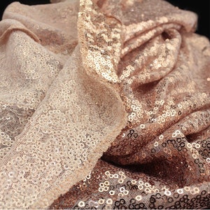 May include: Close-up of rose gold sequin fabric with a sheer mesh backing. The sequins are small and tightly packed, creating a shimmering effect.