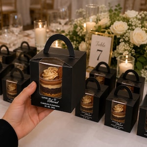 May include: A black square cardboard favour box with a handle and a clear plastic window displaying a layered dessert cup inside. The box features the white printed text "LOVE IS Sweet", "CAROLINE & JOHNSON", and the date "22.02.2032" on the front.