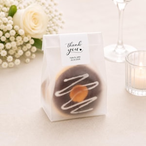 May include: A transparent bag containing a chocolate-frosted doughnut with white icing swirls and a caramel centre. The bag has a white label that reads "thank you" with a heart, the names "Kate & Jake" and the date "20.02.2028".