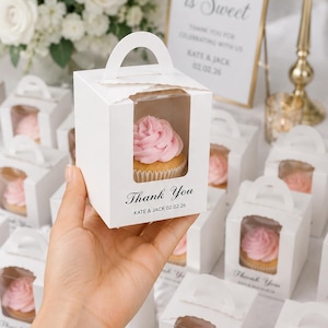 May include: A white cardboard cupcake box with a clear plastic window, a top handle, and the printed text Thank You, KATE & JACK 02.02.26. The box contains a single cupcake with pink frosting. Multiple identical boxes are arranged in the background.