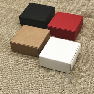 Puede incluir: Cuatro cajas de regalo cuadradas en diferentes colores: marrón, negro, rojo y blanco. Las cajas están hechas de cartón y tienen un diseño sencillo.