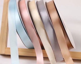 May include: A selection of satin ribbon spools in various pastel colors, including light blue, pink, cream, gray, and tan. The ribbons are displayed on a wooden stand, with the ends of the ribbons cascading down. Ideal for crafts and gift wrapping.
