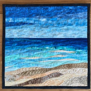 Puede incluir: Un tapiz de pared acolchado que representa un paisaje marino. La obra de arte presenta un degradado de azules que representan el cielo y el océano, con blanco y beige que representan la arena. La pieza está enmarcada con un borde azul oscuro y muestra un acolchado detallado.