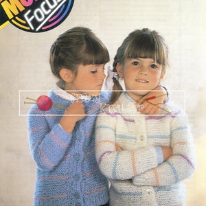 Può includere: Due ragazze indossano cardigan a righe di maglia. Il cardigan a sinistra è blu con strisce rosa. Il cardigan a destra è bianco con strisce rosa, blu e viola. Le ragazze indossano gonne abbinate.