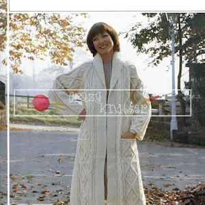 Op de afbeelding: Een witte Aran-gebreide cardigan met een shawlkraag, zakken en een kabelbreipatroon. De cardigan wordt gedragen door een vrouw die in een park staat met herfstbladeren op de grond. De tekst "Aran knitting" en "434" is zichtbaar in de rechterbovenhoek van de afbeelding.