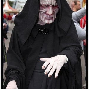 May include: A person in a black hooded robe and mask resembling a character from a science fiction film. The mask is pale with red accents, and the person is wearing white gloves and holding a cane. The costume is likely for a cosplay event.
