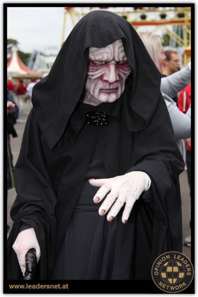 May include: A person in a black hooded robe and mask resembling a character from a science fiction film. The mask is pale with red accents, and the person is wearing white gloves and holding a cane. The costume is likely for a cosplay event.
