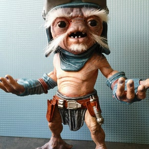 May include: A detailed figurine of a humanoid alien with a wrinkled face, white beard, and goggles. The figure wears a blue and brown outfit with a utility belt and a silver-coloured weapon. The figure is standing with its arms outstretched.
