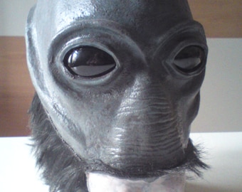 May include: A gray alien mask with large black eyes and a textured, fleshy nose. The mask has black fur around the edges and a pink, fleshy lower jaw.