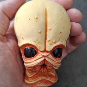 Puede incluir: Una escultura detallada de una cabeza alienígena, sostenida en una mano. La cabeza es principalmente de color crema con acentos naranjas y marrones. Presenta grandes ojos negros y una superficie texturizada.