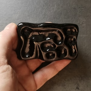 May include: A rectangular, glossy black ceramic object with an abstract design of raised, rounded shapes. The surface reflects light, creating highlights and shadows. The object is held in a hand.