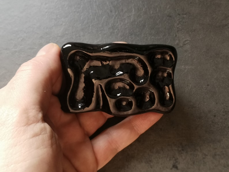 May include: A rectangular, glossy black ceramic object with an abstract design of raised, rounded shapes. The surface reflects light, creating highlights and shadows. The object is held in a hand.