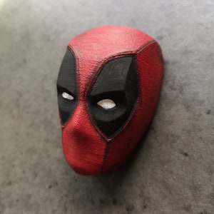 Può includere: Una maschera di Deadpool stampata in 3D di colore rosso e nero con occhi bianchi.