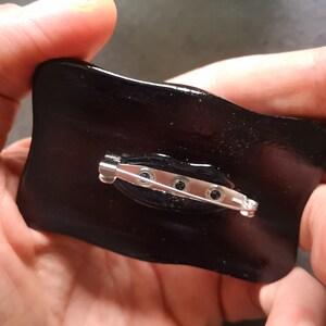 May include: A glossy black pin with a silver safety pin clasp. The pin has a rectangular shape with slightly curved edges. The back of the pin is visible, showing the clasp mechanism and the attachment points.