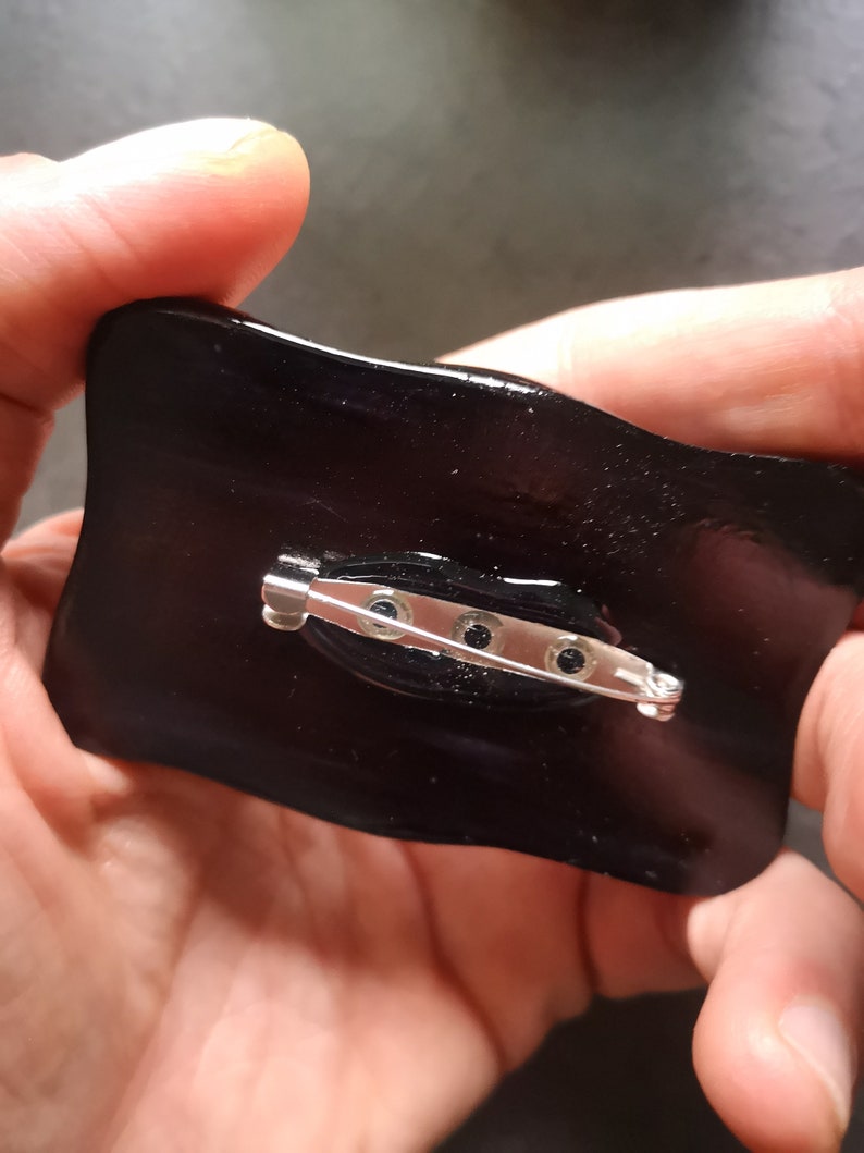 May include: A glossy black pin with a silver safety pin clasp. The pin has a rectangular shape with slightly curved edges. The back of the pin is visible, showing the clasp mechanism and the attachment points.