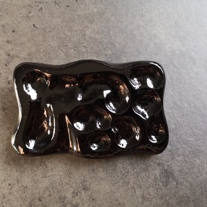 May include: A rectangular, glossy, dark brown ceramic soap dish with an organic, bubbly design. The dish has a smooth, reflective surface and is designed to hold soap. The dish is on a gray surface.