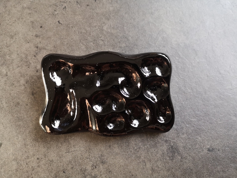 May include: A rectangular, glossy, dark brown ceramic soap dish with an organic, bubbly design. The dish has a smooth, reflective surface and is designed to hold soap. The dish is on a gray surface.
