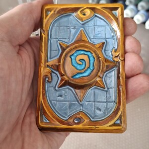 Puede incluir: Una tarjeta rectangular con un diseño detallado. La tarjeta presenta un fondo azul con un diseño de estrella dorada en el centro. Los bordes están enmarcados con un borde dorado. La tarjeta se sostiene en una mano.
