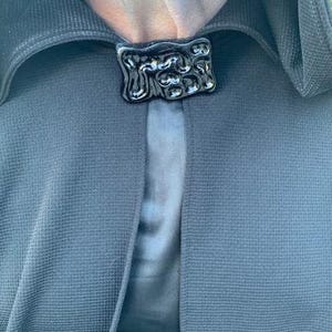 May include: A close-up of a black cloak clasp. The clasp is rectangular with an abstract, textured design. The cloak is a dark blue-grey color, with a textured weave. The background is a lighter blue-grey.